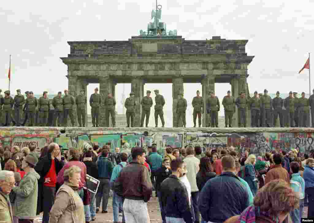 Soldați de gardă din estul Berlinului stând pe bordura de deasupra Zidului Berlinului, în fața Porții Brandenburg, pe 11 noiembrie 1989, la două zile de la &bdquo;prăbușirea&rdquo; Zidului. Cu alte cuvinte, pe 9 Noiembrie 1989, după ce un oficial est-german a anunțat că punctele de trecere ale Zidului vor putea fi traversate de civili fără probleme și &bdquo;fără întârziere&rdquo;, zeci de mii de oameni au trecut în cursul nopții în Berlinul de Vest. Pentru prima dată de când fusese ridicat, Zidul nu a mai fost impenetrabil și a început să fie demolat.&nbsp;
